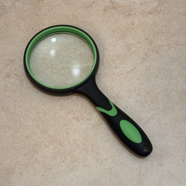 10X shatterproof handheld magnifier with a large round lens rimmed in bright green rubber and a black-and-green rubberised handle, laid flat on a cream limestone counter